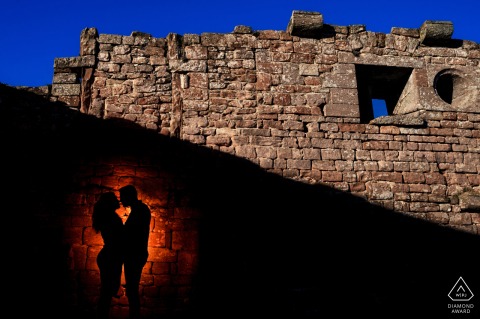Lifestyle couple portrait beautifully composed at Château du Haut Barr - Saverne. Silhouette of a couple in a castle, with a red spotlight accentuating them, near a stacked square stone wall against a deep blue sky.