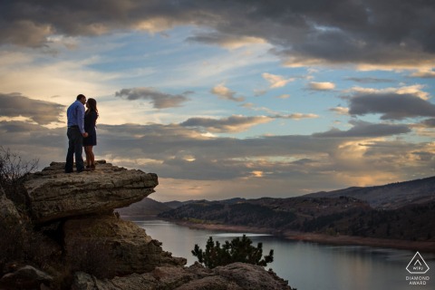 Lifestyle couple portrait at Horsetooth Reservoir, Fort Collins, Colorado featuring a couple perched on a rock, admiring the picturesque sunset and blue hour hues over the serene, still waters below.