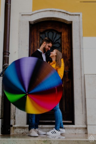 Creative couple portrait by a Székesfehérvár wedding photographer, capturing love in the rain as they stand on the porch of an old building, with the man spinning a rainbow umbrella.