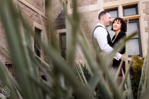 An engaged couple poses in front of Colorado College, sharing a laugh with a soft-focus plant in the foreground, capturing a joyful and candid embrace and look.