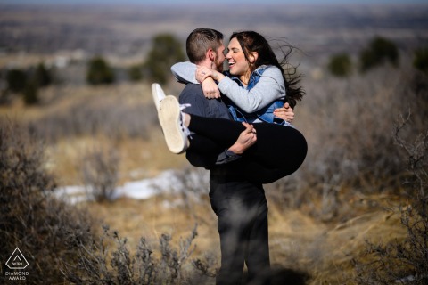 In a beautiful setting at Horsetooth Reservoir, Fort Collins, Colorado, the man sweeps his joyful fiance off her feet, both smiling and having fun during their playful engagement portrait session.