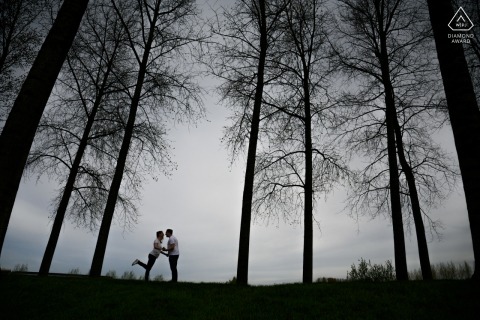 An engaged couple stands close together under a cloudy sky in East Flanders, Flanders. They kiss and hold hands, framed by trees.
