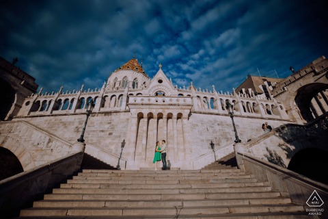 A couple stands elegantly at the top of  set of steps leading to an old-world building in Budapest, Hungary, capturing their engagement portrait against a backdrop of stunning architectural detail.