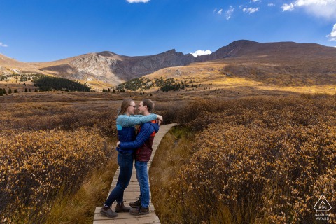 A joyful newly engaged couple embraces in front of a stunning Colorado landscape, having successfully conquered one of the state's famous 14,000-foot mountain peaks. Their love reaches new heights as they bask in the scenic beauty and celebrate.
