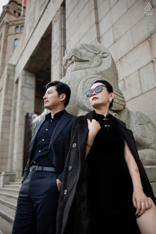 Image of a stylish engaged couple, captured by a wedding photographer at the Bund in Shanghai, China. The couple is standing outside a historic stone building, at the intricately carved stone lion. Both dressed in all black and wearing sunglasses.