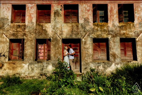 At Caminho de Pedras in Bento Gonçalves, Rio Grande do Sul, an engaged couple stands before a charming two-story building with many windows. Lush green foliage in the foreground adds a touch of nature to the beautiful composition.