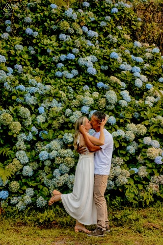In Bento Gonçalves, Rio Grande do Sul, an engaged couple shares a delightful kiss, with her playfully lifting a foot. A stunning wall of summer flowers blooms behind them, perfectly balancing the scene.