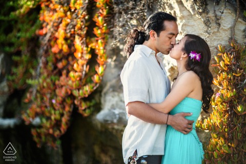 A couple shares a romantic kiss on a sunlit beach in Santa Cruz, CA, surrounded by coastal foliage, capturing the warmth and beauty of Northern California.