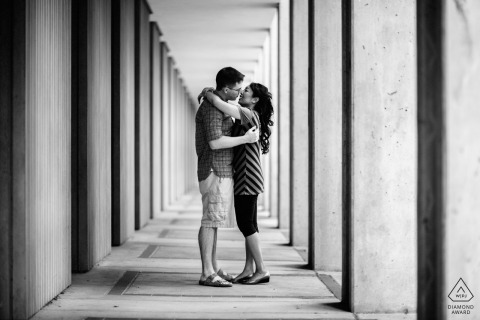 In the grand hallway of a veranda at UC Santa Cruz, a couple embraces in a romantic black-and-white kiss, framed symmetrically by repeating architectural lines.