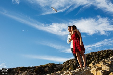 An engaged couple stands lovingly together at Natural Bridges State Beach in Santa Cruz, CA, perfectly silhouetted against the expansive blue sky and fluffy white clouds.