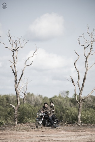 In Chantaburi, Thailand, a couple poses on a motorbike in a tall vertical image, flanked by towering dead trees. This striking portrait is expertly captured by a skilled WPJA photographer.