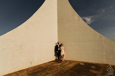 A couple from Brasilia poses cheerfully in the bright sunlight, standing at the corner of an artistic backdrop. 