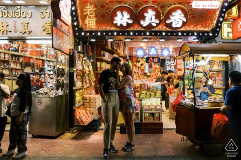 In Bangkok's Chinatown, the couple shares a kiss in front of a bustling, traditional Chinese shop, vibrant and lively.
