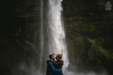 Engaged couple shares a kiss with a majestic waterfall cascading in the background on Iceland's stunning south coast.