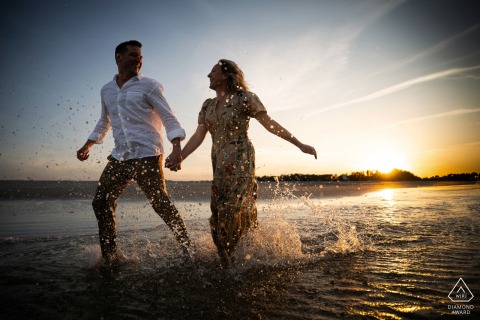 A couple joyfully runs through shallow water at Crane Beach, Ipswich, MA, during golden hour, creating romantic reflections.
