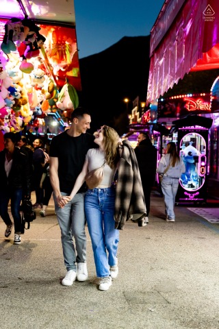 Lovers stroll hand-in-hand through the lively aisles of Grenoble's Foire des Rameaux, surrounded by vibrant fairground attractions.