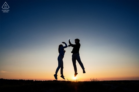A couple joyfully jumps atop the Wassenaar dunes at sunset, silhouetted against a vibrant sky, embracing happiness.