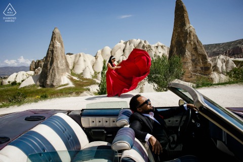 Bride in flowing red dress poses beside convertible car in Goreme’s Cappadocia desert, engagement session.