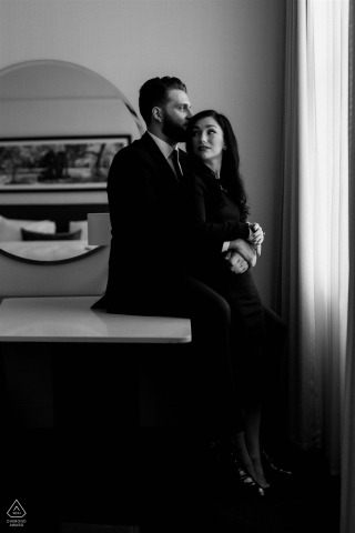 Elegantly Dressed Couple Sits by Loews Hotel Window, Romantic Philadelphia Engagement Portrait A couple elegantly dressed sits by a hotel room window at Loews Hotel, Philadelphia, showcasing a romantic engagement portrait.