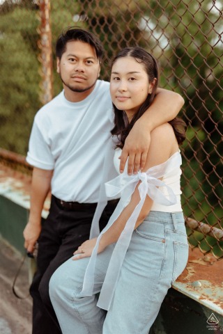 Engaged couple posing on a walking bridge above Lincoln Highway, chain link fence backdrop, Philadelphia, PA.