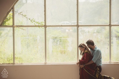 Couple in Chennai, India embracing indoors; he hugs her from behind near large clear window panes, natural daylight illuminating them.