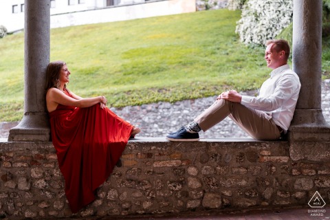 Couple in Udine, Italy, seated on castle wall, facing each other with backs against separate pillars, sharing a quiet session.