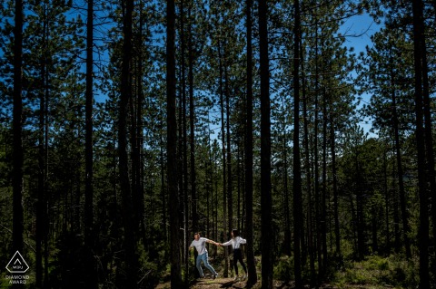 Pre-wedding couple holding hands, playfully walking along the edge of tall, dark forest trees in Sierra de las Villas, Jaén, Spain.