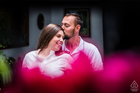 Pre-wedding couple in Cambil, Jaén, Spain; man stands behind kissing her head, soft focus pink florals foreground.