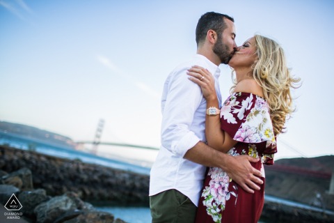 Engaged couple kisses in Sausalito, California, with iconic bridge and sparkling water in the background.