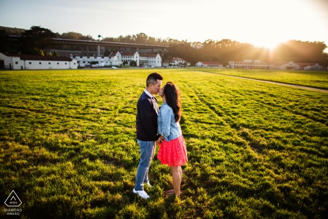 In Northern San Francisco, California, a couple shares a kiss in an open grass field, with the low sun casting warm tones. This engagement portrait is captured by an expert WPJA photographer.
