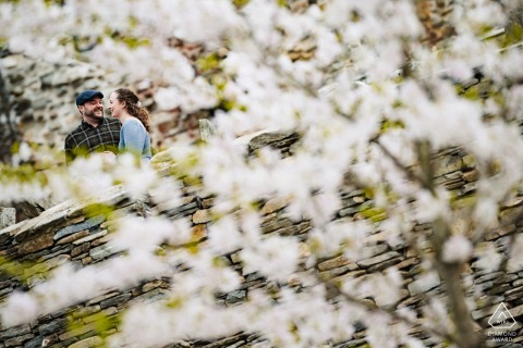 At Gillette Castle State Park, Connecticut, a couple's engagement is enchantingly captured through blooming white flowers, revealing their connection amidst the park's historic charm and natural beauty by a WPJA pro photographer.