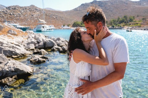 At Agia Marina Beach, Symi, Greece, a couple shares a tender kiss with the turquoise sea as their stunning backdrop, beautifully captured by a WPJA pro photographer.