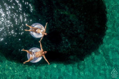 High above the crystal-clear waters of Agia Marina Beach in Symi, Greece, a drone captures a couple on swim rings, holding hands. This enchanting pre-wedding portrait by a skilled WPJA photographer beautifully symbolizes their unity and adventure.
