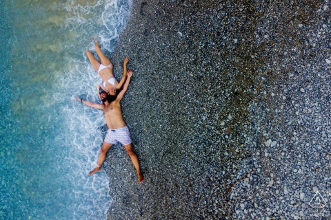 At Agios Georgios Dysalona Beach in Symi, Greece, a drone captures a breathtaking scene as a couple lies neck to neck on the shoreline, embraced gently by waves. This striking pre-wedding portrait, masterfully taken by a WPJA photographer.