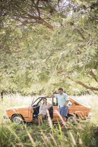 Couple poses with car in a grassy field under a tree in Chantaburi, Thailand, smiling warmly.