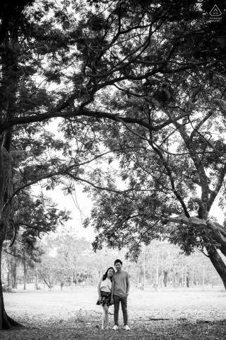Black and white vertical portrait of couple standing side by side under trees in Bangkok, Thailand.
