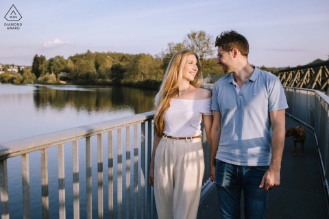 Couple walks along railed pathway at Biggesee lake, Germany, enjoying sunset in a tranquil scene.