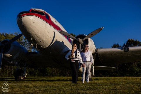 Engaged couple poses confidently under vintage aircraft at Cape Cod Airfield, radiating cinematic modern romance.