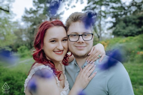 A joyful couple embraces in lush Gellért-hegy park, framed by soft-focus purple foreground flowers.
