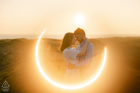 Engaged couple stands on sand dune, backlit by fiery ring and sunset, overlooking Les Sables d'Olonne ocean.
