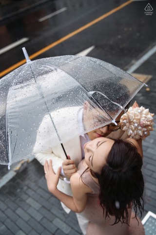Engaged couple embraces under clear umbrella on rainy Bund pavement, Shanghai, for a sweet portrait.