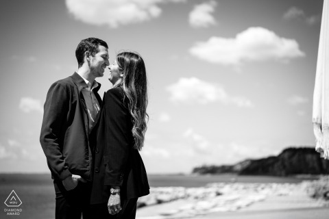 Laughing couple face-to-face on Portopiccolo shoreline, black and white, waves’ white water rolling in.