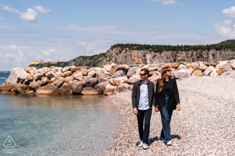 Couple strolls along rocky seashore in Portopiccolo, portrait with calm coastline beside them.