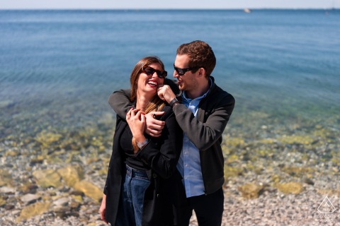 Couple stands together on Portopiccolo seashore, romantic portrait with sea and rocky shore in background.