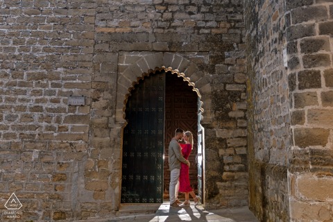 Pre-wedding portrait: The couple stand near archway of historic brick building in Baeza.