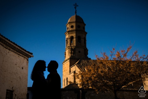 Silhouetted couple poses against deep blue sky, historic architecture towering behind, Baeza, Jaén, Spain.