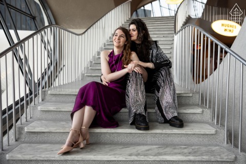 Couple sits on TWA Hotel steps, gazing outside at JFK Airport tarmac through large glass windows.