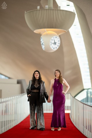 Couple stands stoically, hand in hand, beneath retro clock atop TWA Hotel, JFK Airport, NY.