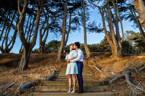 Couple stands on outdoor San Francisco stairway, surrounded by tall trees, embracing in natural light.