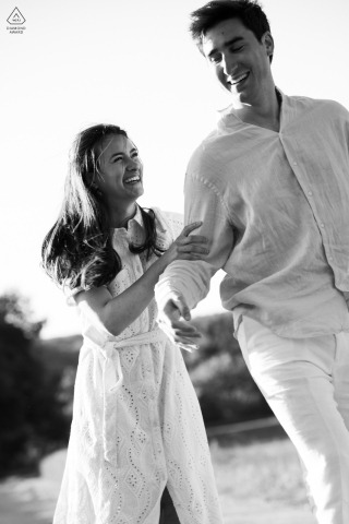 Grignan, France. A black and white, vertical engagement portrait of a couple walking and laughing.
